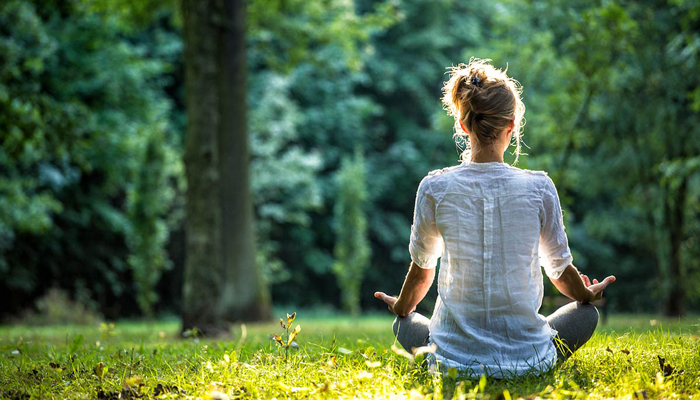 Vrouw in wit shirt zit te mediteren op een grasveld in de zon met haar rug naar de kijker toe en haar handpalmen open op haar knieën