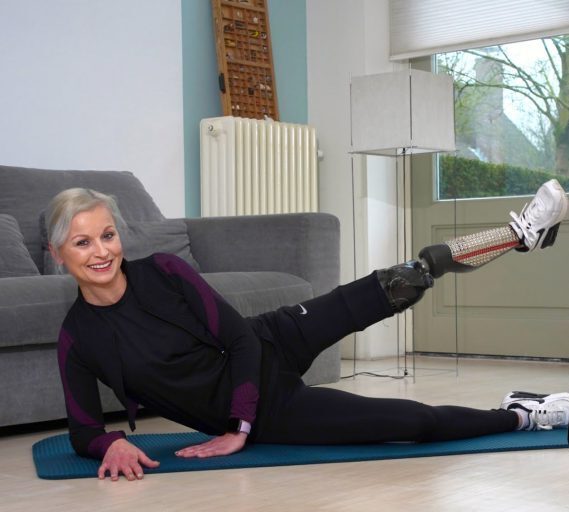 vrouw in zwart trainingspak doet beenoefening op yogamat voor de bank met plaatje van Uniek Sporten app