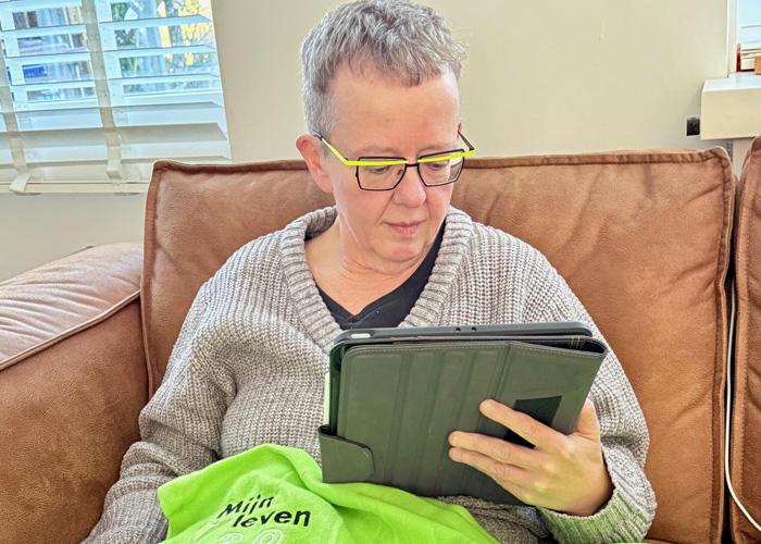 Vrouw met kort grijs haar en een bril met een felle groene bovenrand op de bank met een iPad onder een groen fleecedekentje