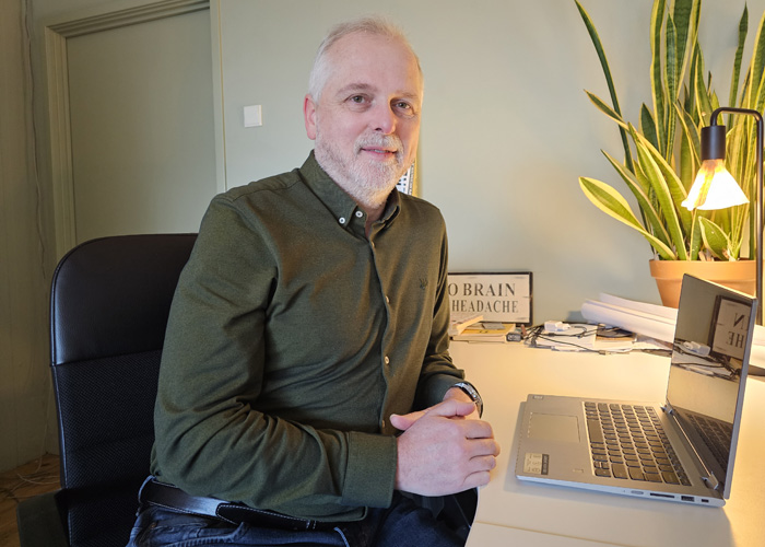 man met olijfgroen overhemd, licht kalend en grijs sikje, aan bureau met laptop, migraine